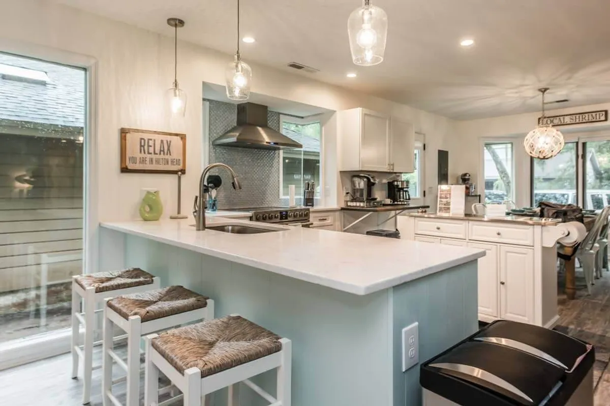 A modern kitchen island with stools sits at the heart of an open-concept kitchen. The space is enhanced by elegant lighting fixtures, visible kitchen appliances, and a window view, creating an atmosphere suitable for both casual brunches and festive gatherings.