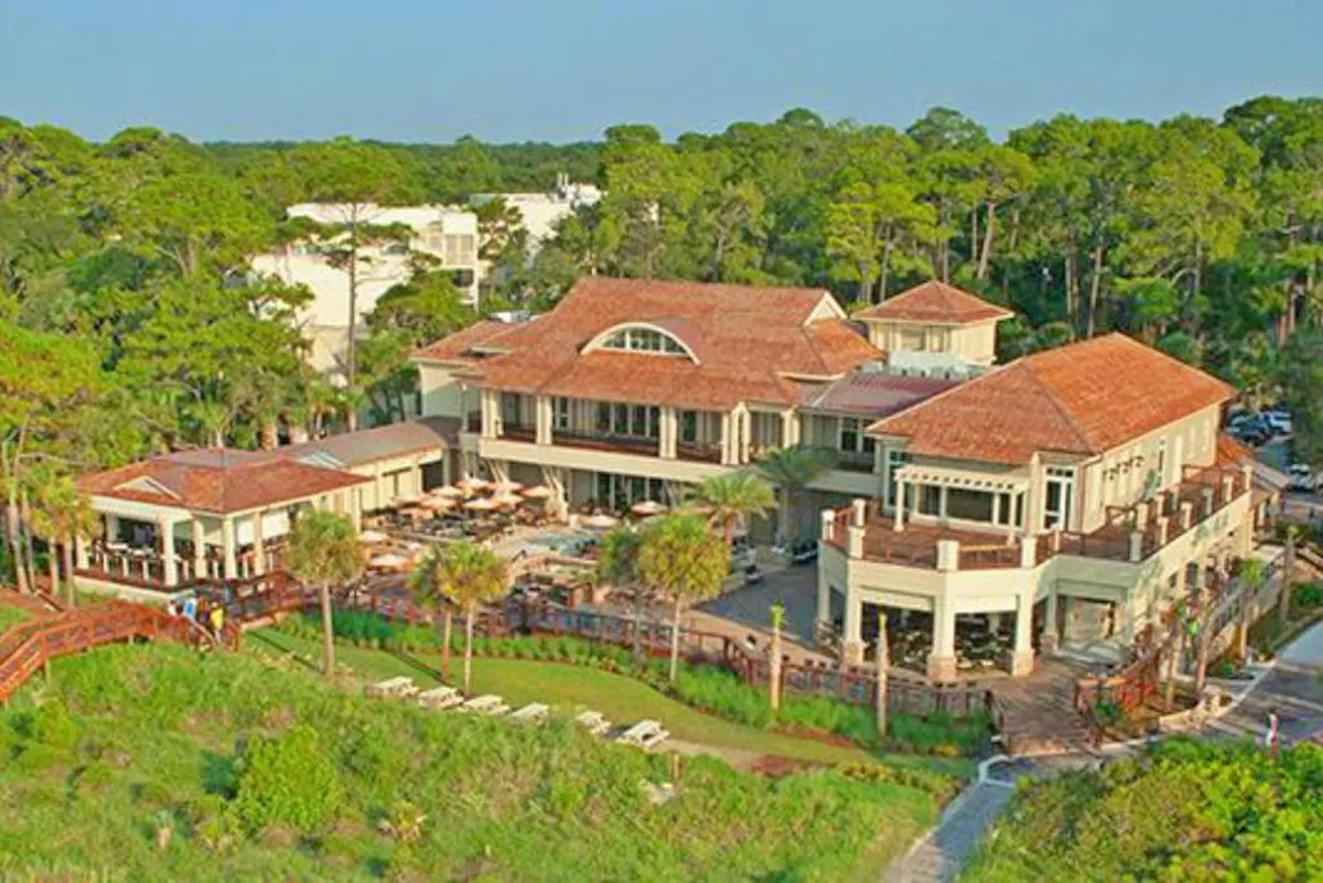 An elegant exterior view of a large central building with outdoor seating areas, surrounded by greenery and a wooden boardwalk. The upscale ambiance and communal spaces hint at boutique shopping, fine dining, and special events—mirroring the vibe of Harbour Town or South Beach Marina.