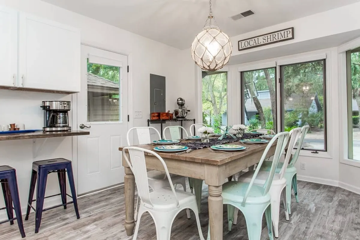 A bright dining area with a central table and chairs, pendant lighting, a coffee maker, and a whimsical 'local shrimp' sign. The room exudes warmth and encourages communal meals, coffee breaks, and game nights, highlighted by its inviting and lively atmosphere.