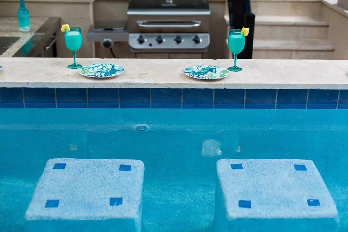 A close-up of two turquoise drinks with lemon garnishes placed on decorative plates, set against the backdrop of a poolside grill and inviting poolside seating. The scene conveys a sense of leisurely poolside lounging and friendly, festive gatherings.
