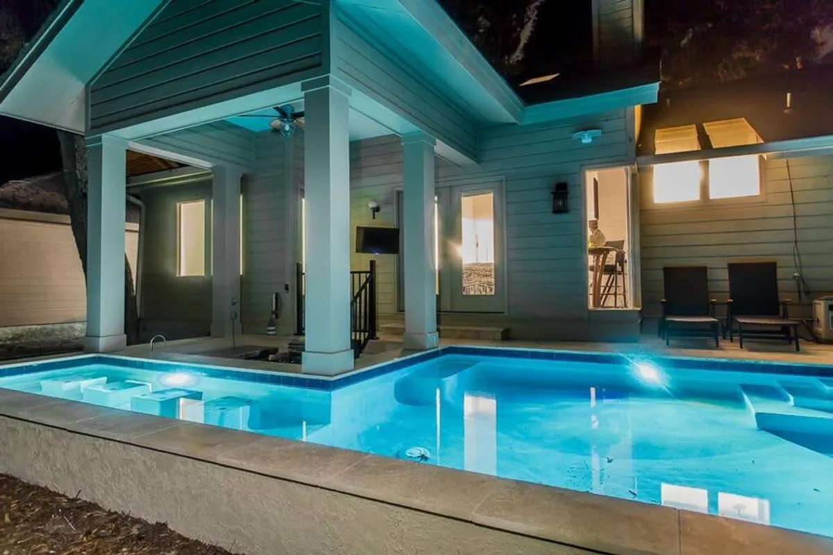 The illuminated pool glows at twilight against a backdrop of modern architecture and an inviting outdoor seating area. The evening setting is sophisticated and serene, perfect for relaxing swims, laughter, and intimate gatherings after sunset.