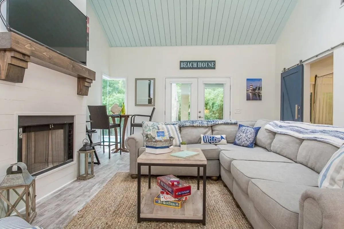 A spacious living room featuring a large sectional sofa, a cozy fireplace, coffee table, and a 'beach house' sign. The area is flooded with natural light and offers plush seating ideal for group gatherings or relaxed lounging. The decor is contemporary coastal, contributing to the sophisticated yet laid-back atmosphere.
