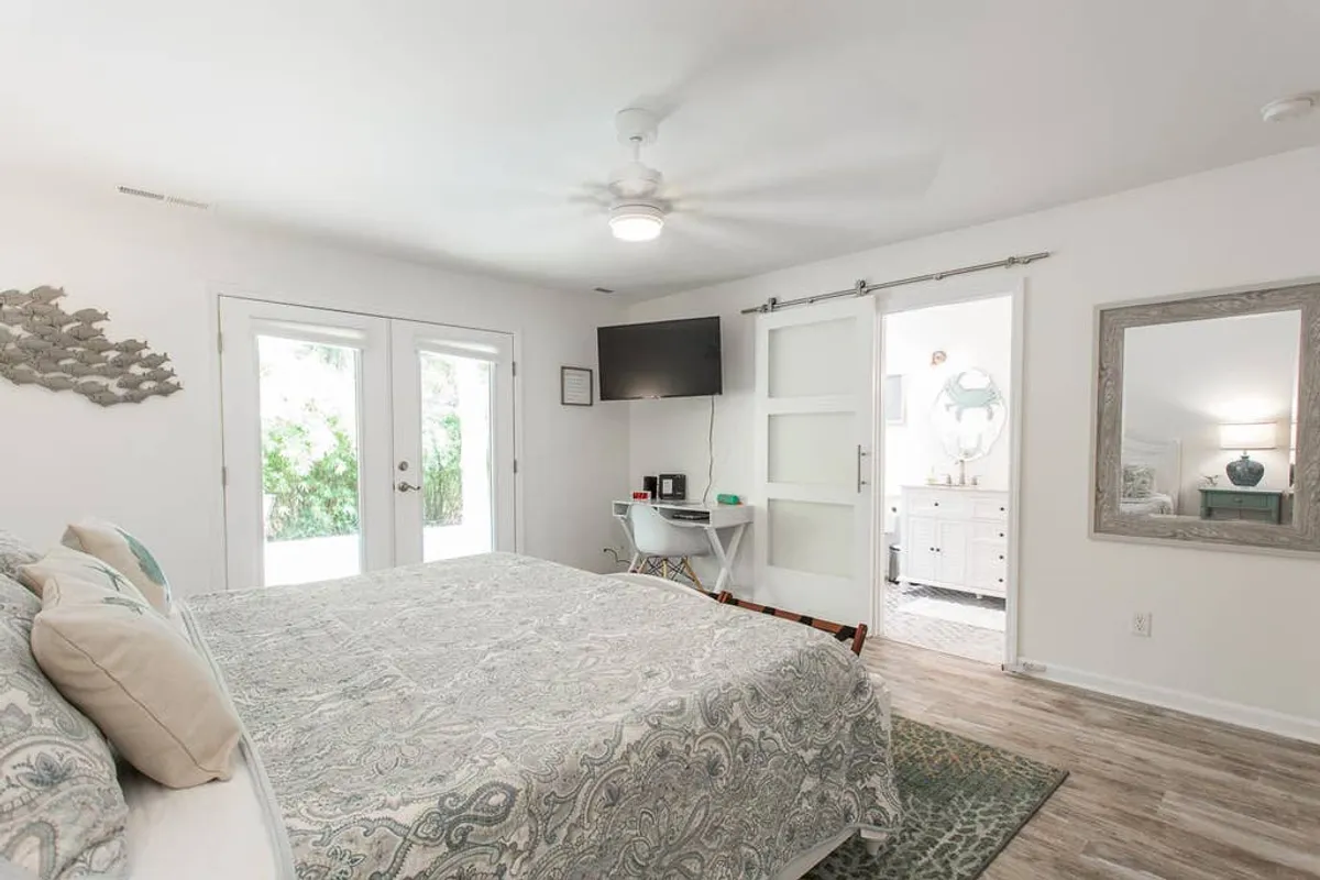 A serene bedroom featuring a plush bed, double doors, a television, and a mirror, bathed in natural light. The ambiance is calm and comfortable, inviting guests to unwind and enjoy a restful night or a cozy movie evening.