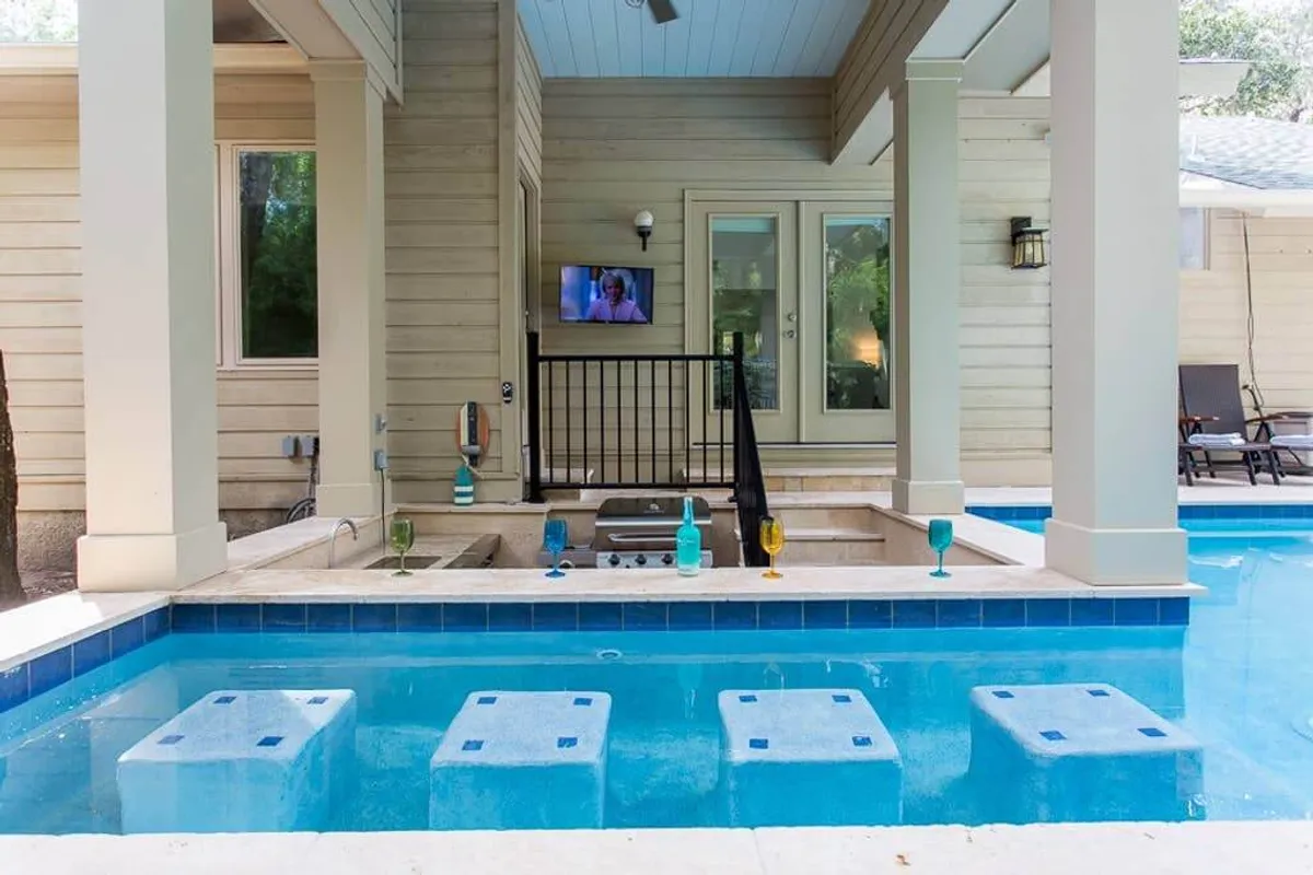This image showcases a sunlit, private pool area featuring a luxurious swim-up bar with submerged bar stools, a wall-mounted television perfect for outdoor movie nights, and inviting spaces for lounging and entertaining. The atmosphere is casual, relaxing, and designed for fun gatherings with friends and family.